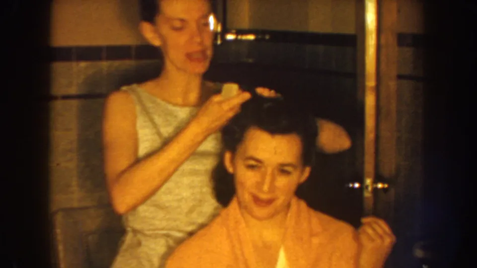 Near the end of the 1959 Nebraska clip, the standing woman smooths the seated woman's hair in a warm, tiled bathroom interior.