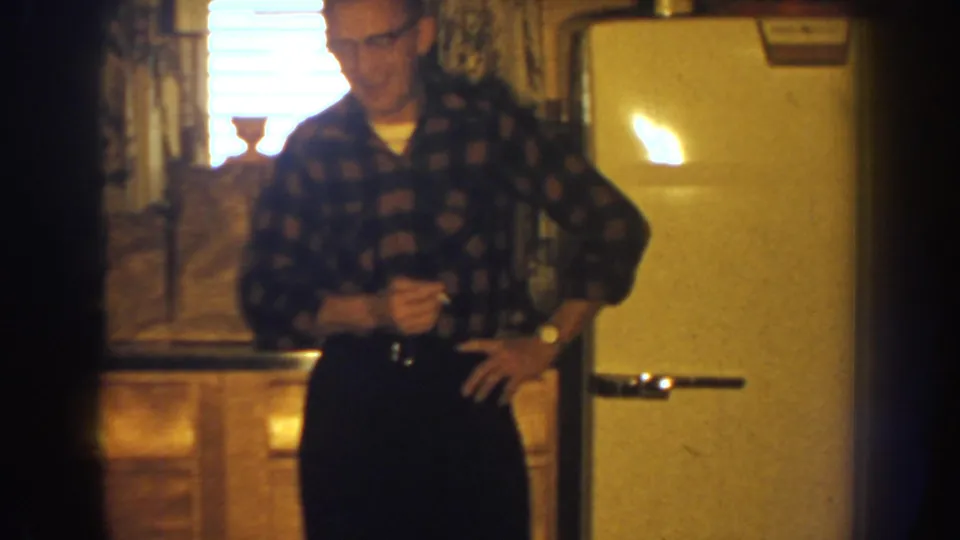 A man in glasses and a dark patterned shirt stands in a home interior near a rounded refrigerator, bright window, and patterned curtains.