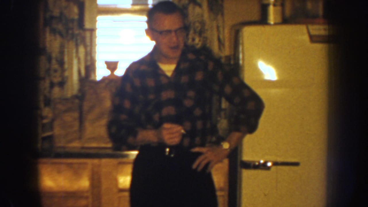 Man in glasses stands in a kitchen beside a cream refrigerator, holding a cigarette and glass.