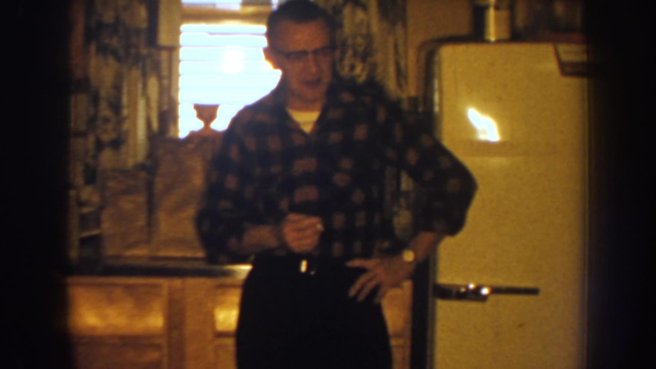 1959 Nebraska kitchen scene with a man in glasses holding a cigarette and glass beside a cream-colored refrigerator.