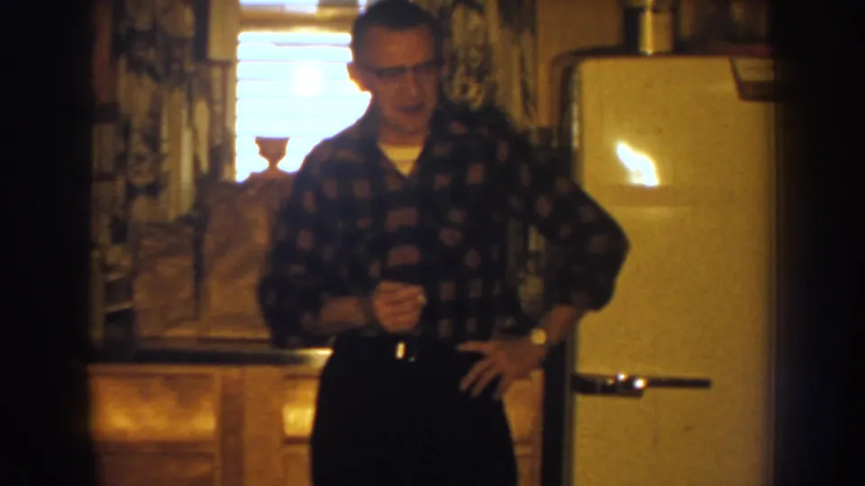 1959 Nebraska kitchen scene with a man in glasses holding a cigarette and glass beside a cream-colored refrigerator.