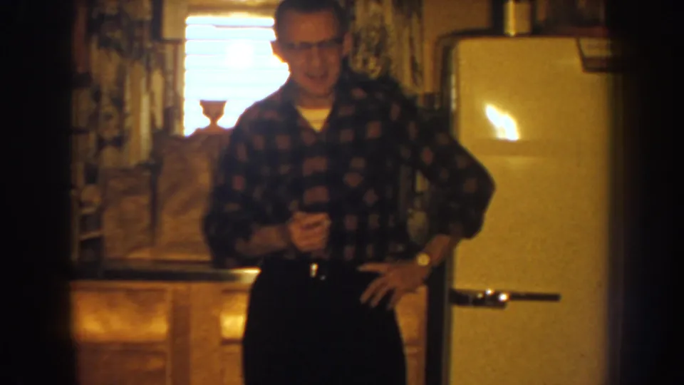 Warm 8mm frame from a 1959 Nebraska kitchen showing a man in glasses beside wood cabinets and a cream refrigerator.