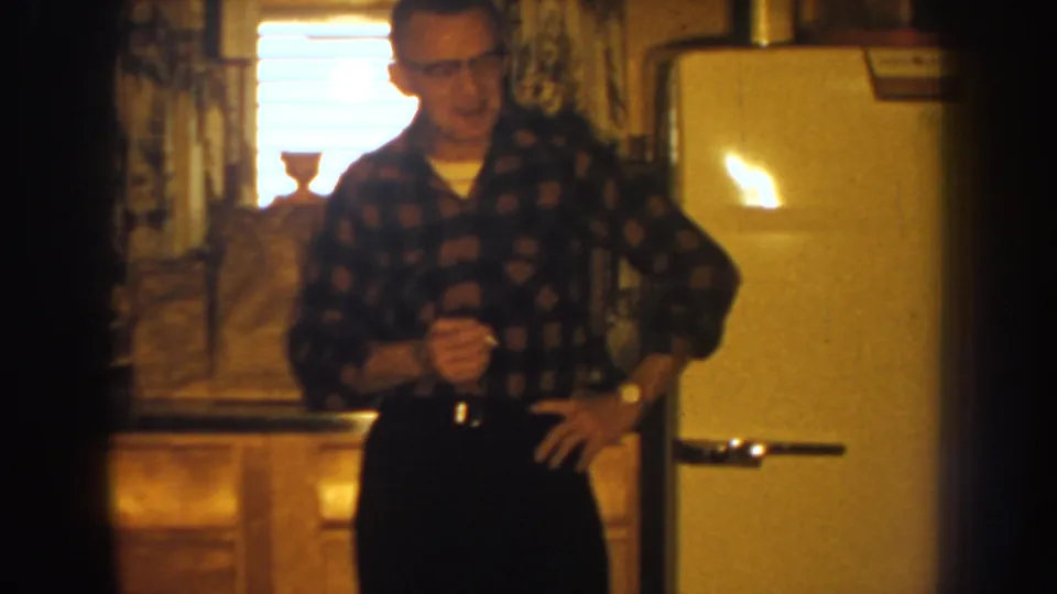 1959 Nebraska home-movie still of a man in a plaid shirt and glasses standing in a private kitchen by the refrigerator.