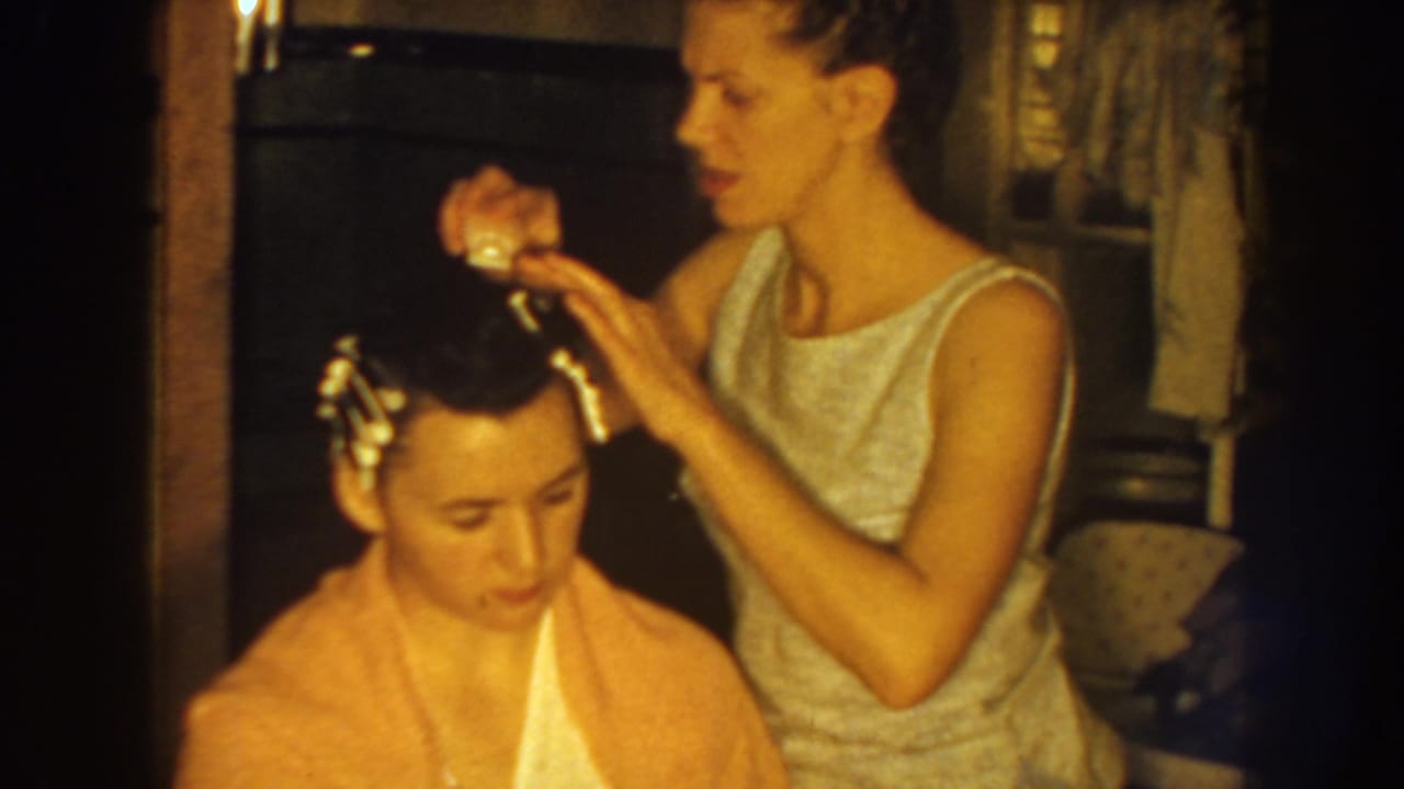 Woman places curlers or clips in a seated boy's hair inside a warm home interior.
