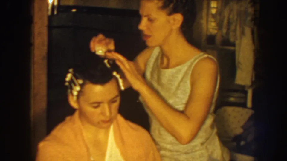 In 1959 Nebraska home-movie footage, a woman stands beside a seated boy and secures curlers or clips in his hair.