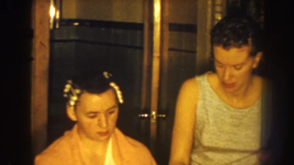 The seated boy faces downward with clips or curlers visible while the woman stands beside him in the warmly lit 1959 Nebraska home scene.