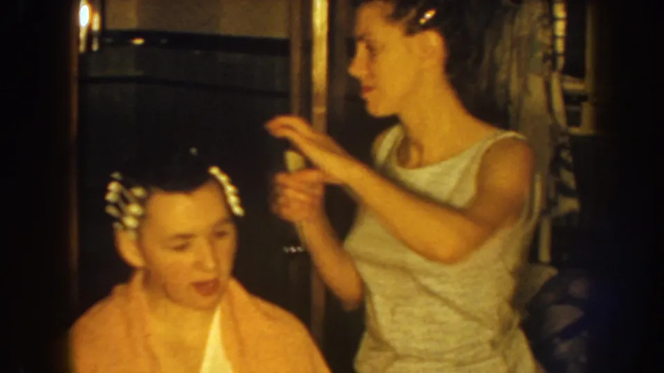 A woman in a sleeveless dress holds a section of the boy's hair as he sits with clips or curlers in a private Nebraska interior.