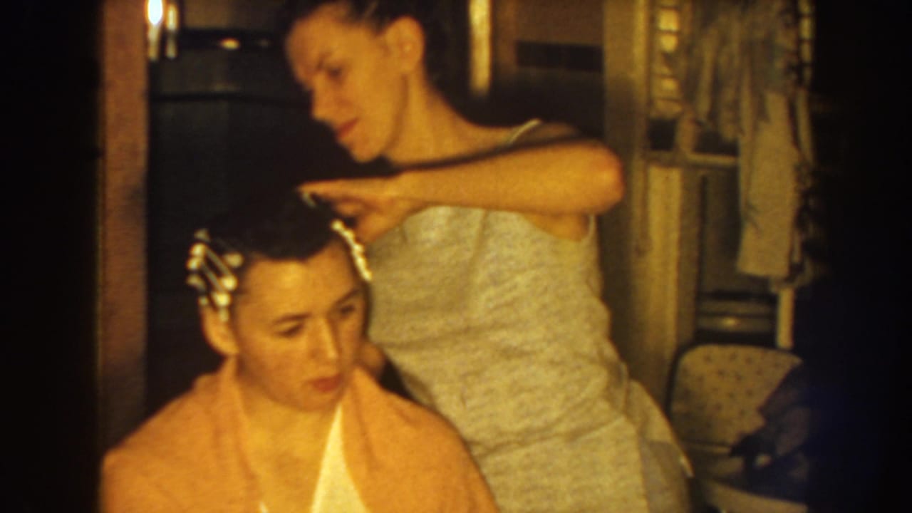 Mother adjusts a seated young woman's hair with rollers in a warm interior.