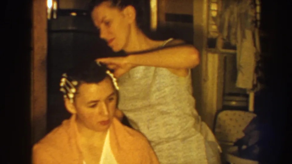 In 1959 Nebraska home-movie footage, a mother stands behind her seated daughter and arranges hair rollers as the daughter wears a yellow towel.