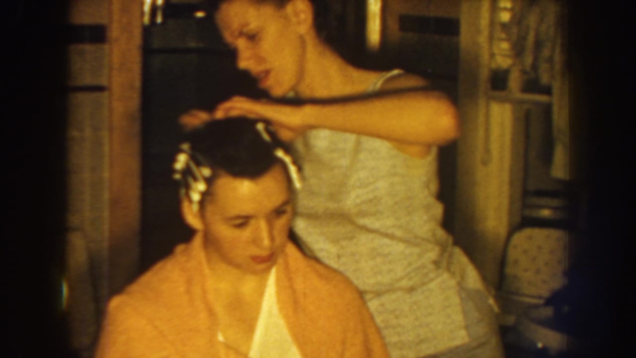 The mother leans over her seated daughter, smoothing dark hair and rollers in a warmly lit Nebraska interior from 1959.