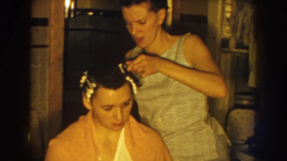 A mother continues arranging her daughter's rolled hair in a private home interior, with warm 8mm color and soft focus.