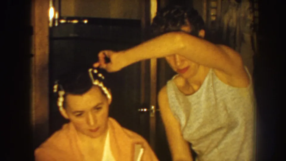 A home-movie frame from 1959 Nebraska shows the stylist reaching across the client's head to adjust white rollers.