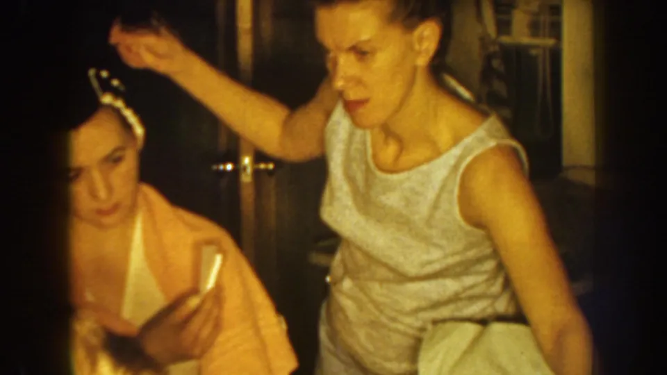 In a Nebraska 1959 home interior, an older sister begins arranging a younger sister's dark hair as she sits wrapped in a yellow towel.