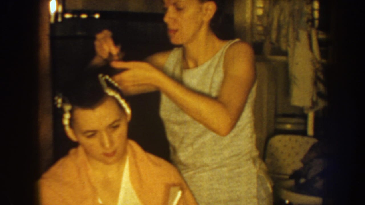 The older sister works carefully with rollers along the younger sister's hairline in a warmly lit 1959 Nebraska home-movie frame.