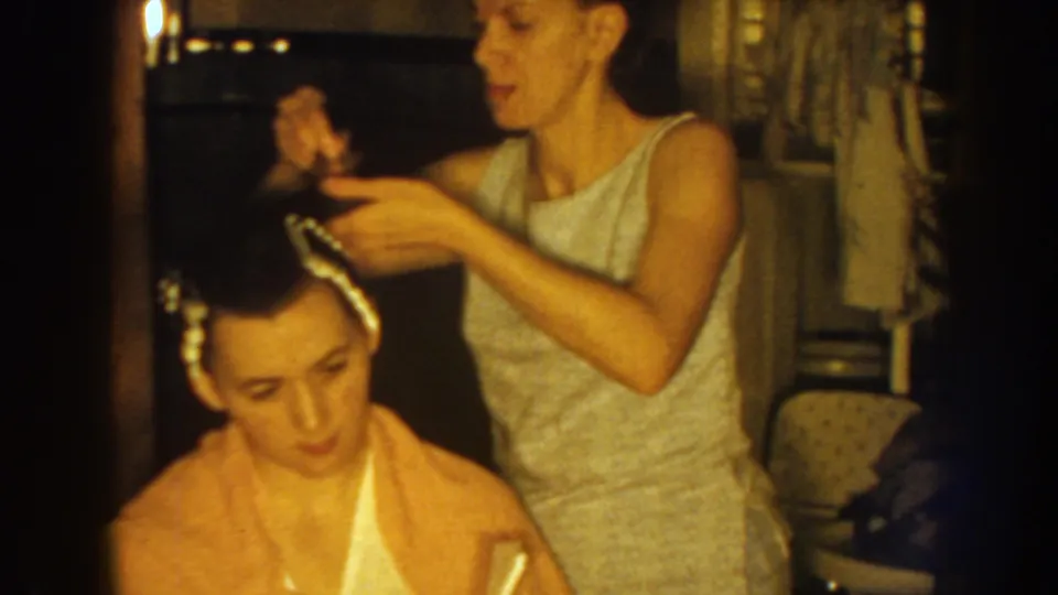 The older sister works carefully with rollers along the younger sister's hairline in a warmly lit 1959 Nebraska home-movie frame.