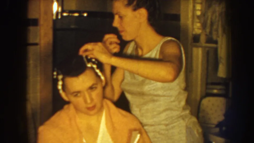 Near the end of the 1959 Nebraska clip, the older sister continues styling the younger sister's hair as the seated girl looks down.