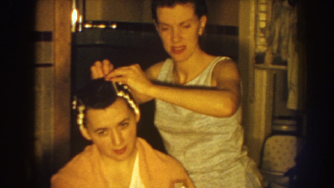 The standing woman continues setting hair rollers on the seated woman in a warm, softly focused 1959 Nebraska home-movie frame.