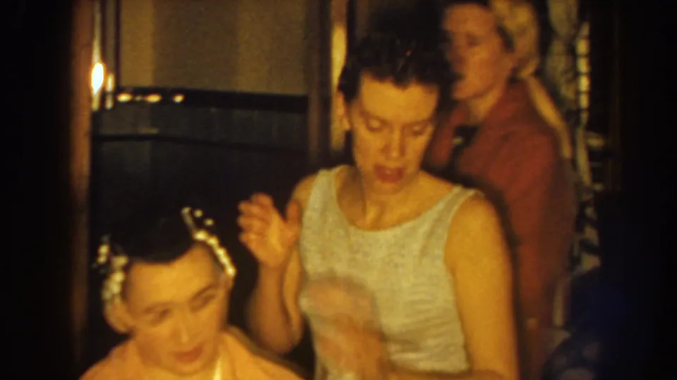 A woman in a sleeveless dress pauses or gestures beside the seated woman with curlers, while another adult appears in the room behind them.