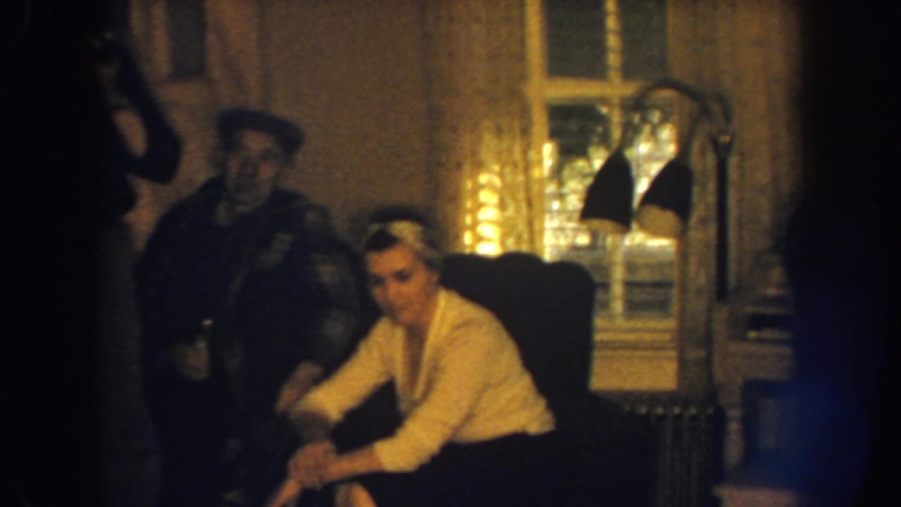 Woman seated in a dark armchair with two men nearby in a dim living room.