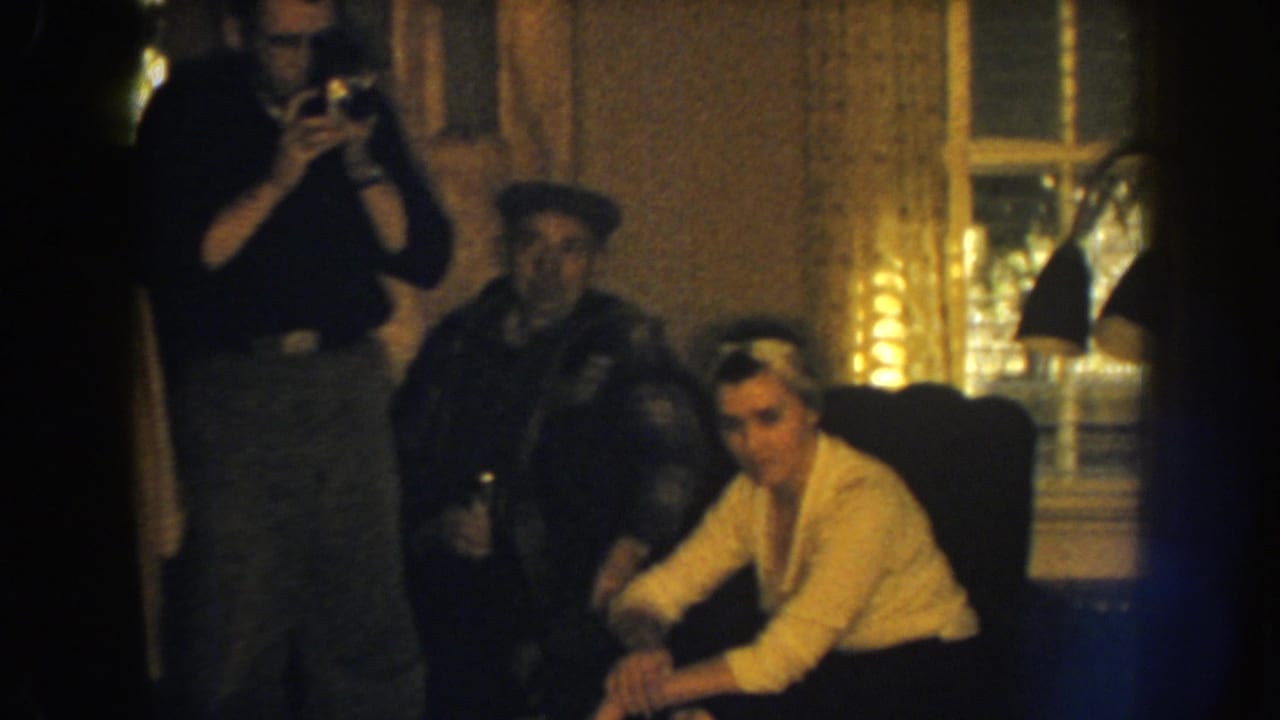 1959 Nebraska interior frame with a man using a camera, a seated woman in a light sweater, and another man in a cap nearby.
