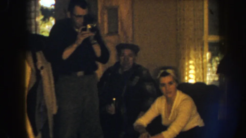 1959 Nebraska home-movie frame showing a camera-holding man beside a seated woman and another man in a dim living room.
