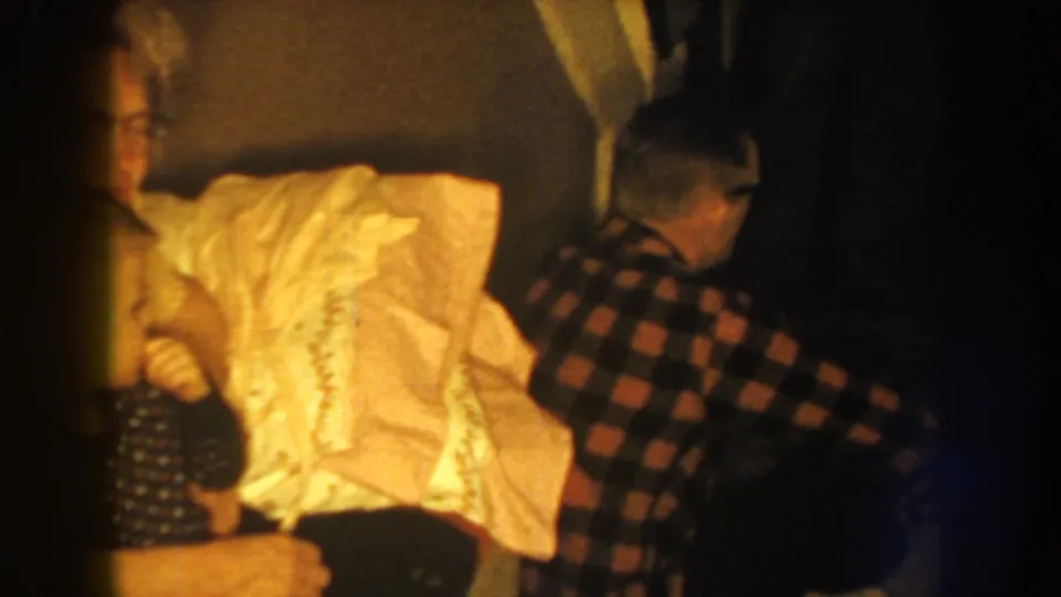 1959 Nebraska home-movie frame with a man in a plaid shirt near a person in light clothing inside a dim private room.