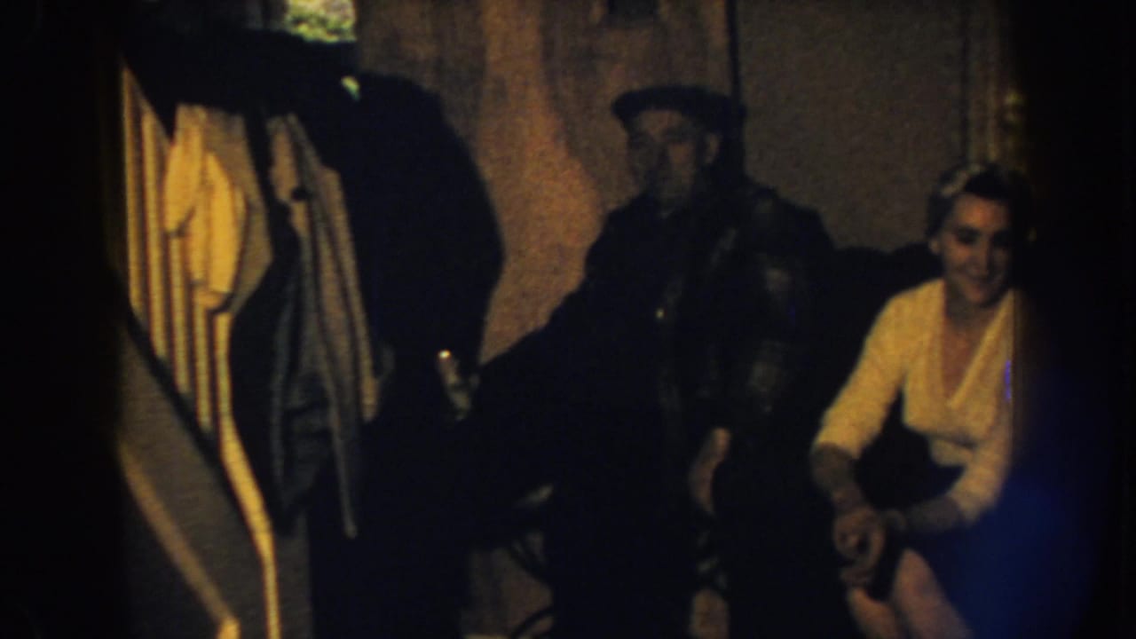 Man in a dark cap stands near a seated woman on a brown sofa in a dim room.