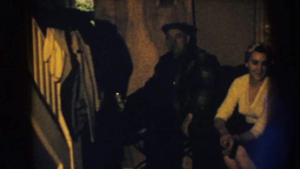 1959 Nebraska home-movie frame of a man in a dark cap standing beside a woman seated on a brown sofa in a low-lit interior.
