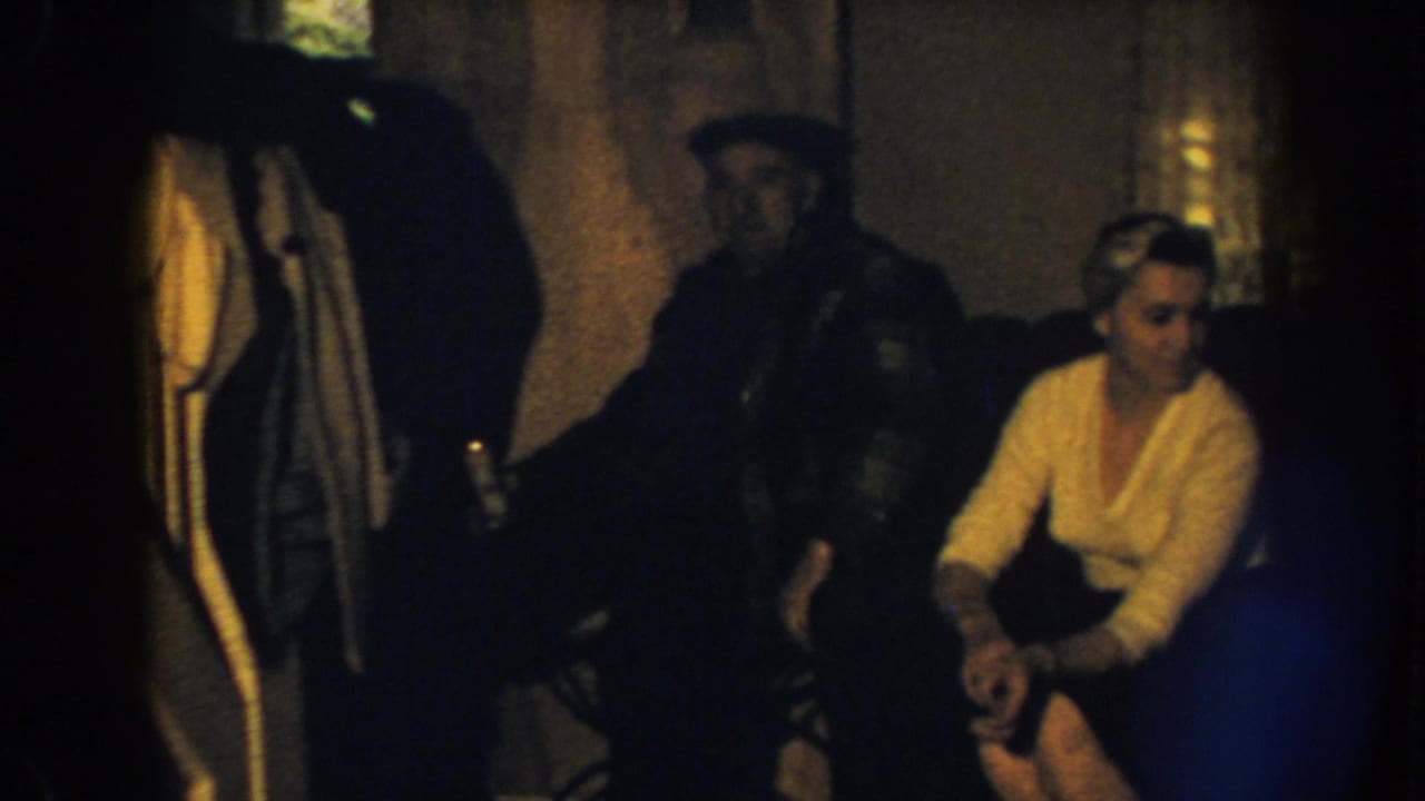 1959 Nebraska home-movie frame showing a woman seated on a sofa with hands together as a man in a cap stands behind her.