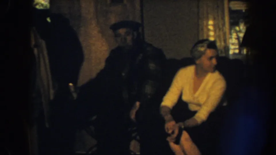1959 Nebraska home-movie frame of a woman on a sofa turning toward a man in a dark cap, with hanging clothes nearby.