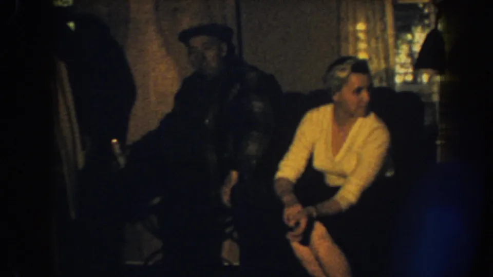 1959 Nebraska home-movie frame showing a woman in a cream sweater looking off screen while a man stands behind the sofa.