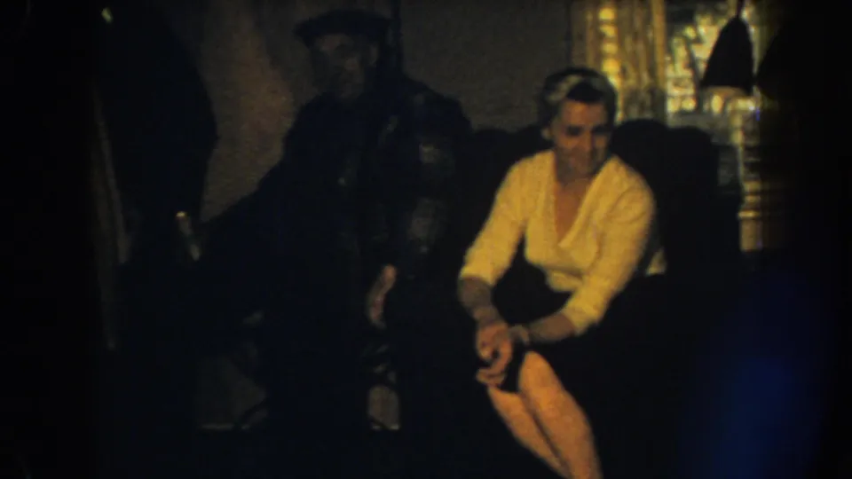 1959 Nebraska home-movie still showing a woman on a couch turning toward someone off-camera, with a man partly hidden behind her.