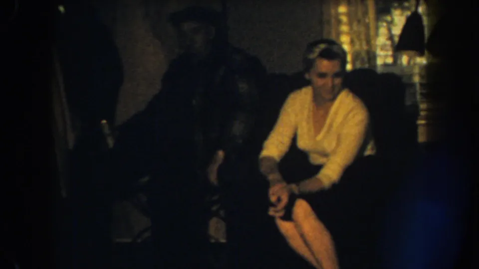 1959 Nebraska 8mm frame of a woman seated on a couch, hands near her knee, with a shadowed man and curtained window behind.