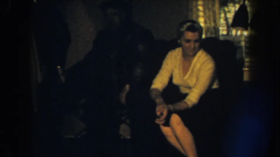 1959 Nebraska home-movie frame with a woman seated on a couch, looking downward, and a man mostly obscured in the background.