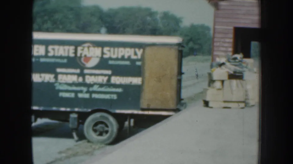 1959 Nebraska 8mm frame with the farm supply truck partly visible near stacked boxes and a red-sided building opening.
