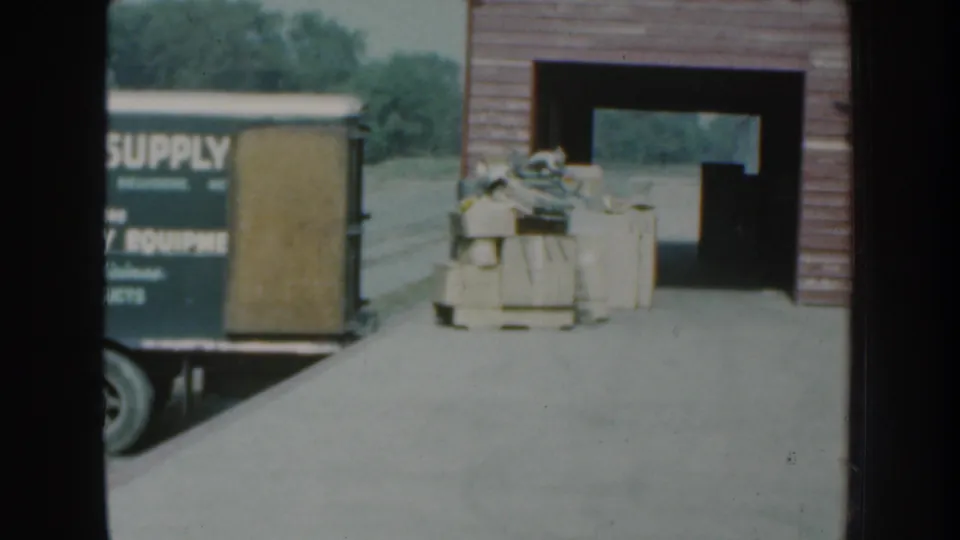 1959 Nebraska 8mm frame with stacked supplies beside an open rural building and the farm supply truck mostly out of view.