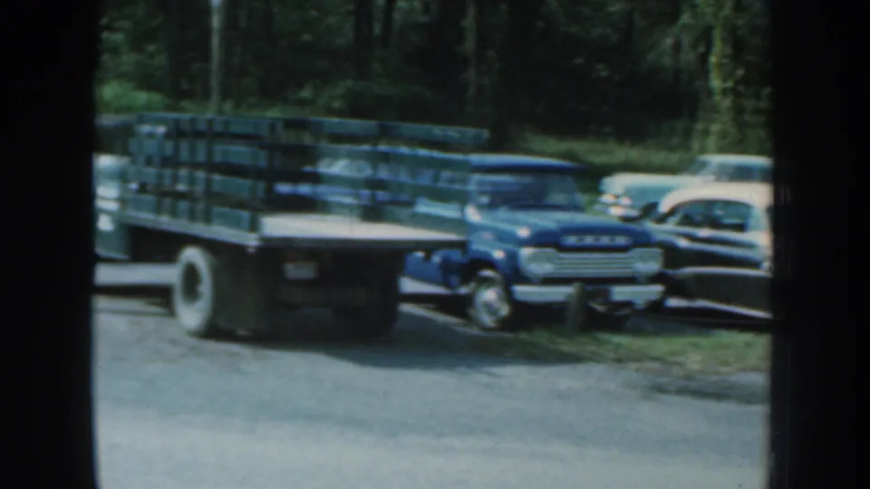 1959 Nebraska frame with a blue pickup truck and several mid-century cars parked near trees.