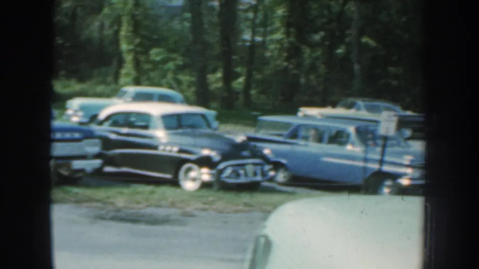 1959 Nebraska 8mm frame showing rounded sedans and a blue station wagon or car parked in a wooded lot.