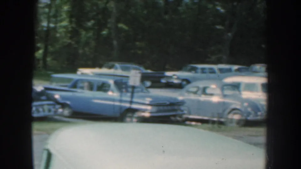 Nebraska 1959 home-movie frame showing pale late-1950s cars parked in a wooded lot, seen through dark window framing.