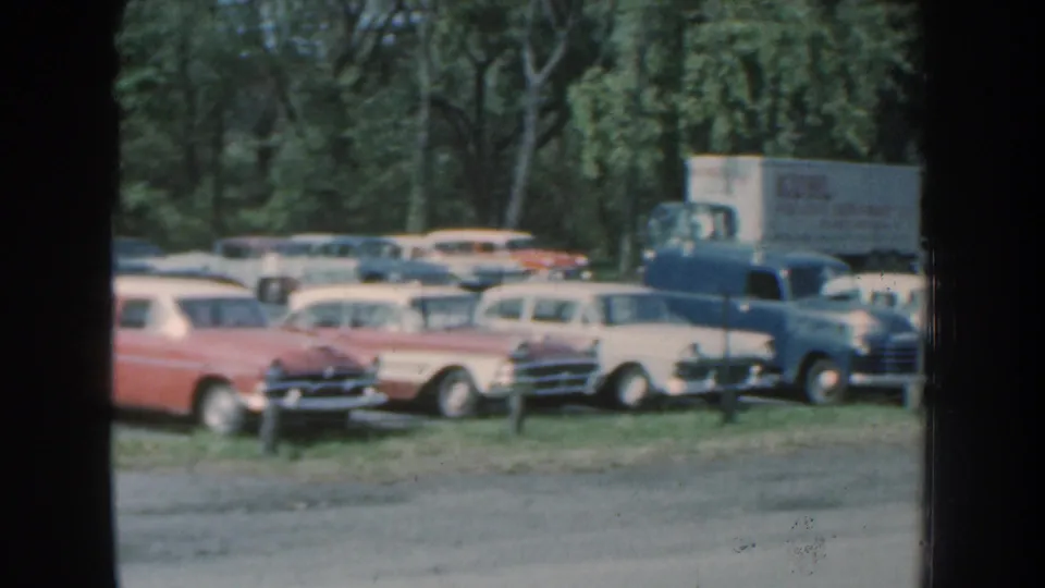 Nebraska 1959 home-movie frame showing a red-and-white wagon, pale sedans, a blue vehicle, and a box truck in a crowded wooded lot.