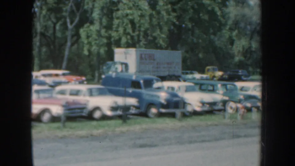 Nebraska 1959 frame with a box truck, pale wagons and sedans, and a dark car parked beside trees in a sunlit lot.