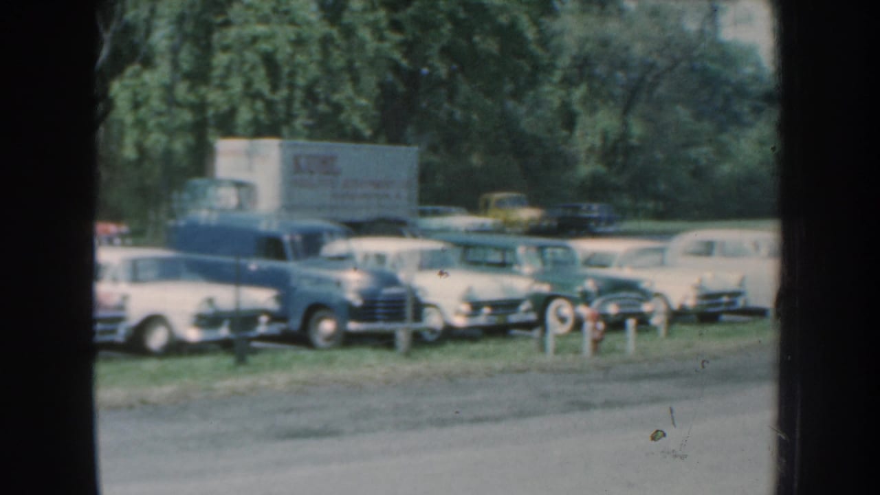 1959 Nebraska 8mm frame showing vintage cars and a light-colored truck parked near a grassy roadside lot.