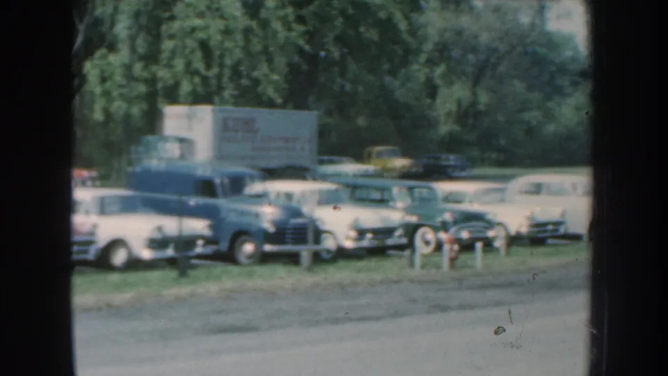 1959 Nebraska 8mm frame showing vintage cars and a light-colored truck parked near a grassy roadside lot.