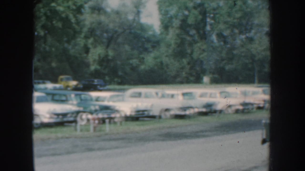 1959 Nebraska roadside view of parked mid-century cars and station wagons behind a road, partly framed by film gate edges.
