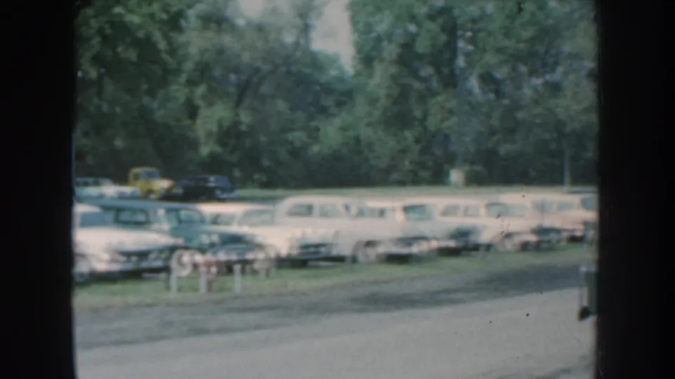 1959 Nebraska roadside view of parked mid-century cars and station wagons behind a road, partly framed by film gate edges.