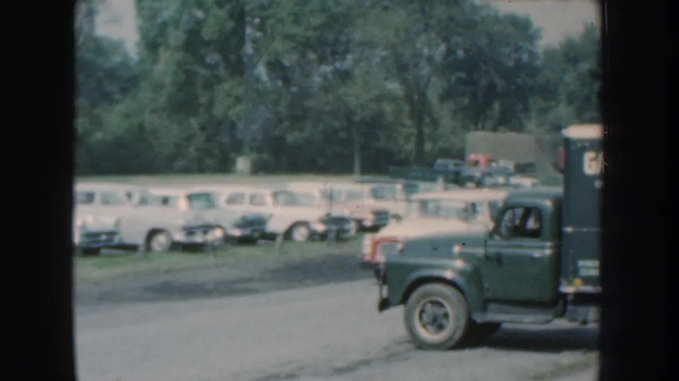 1959 Nebraska 8mm frame with a dark pickup and box truck moving past a lot of parked vintage cars.