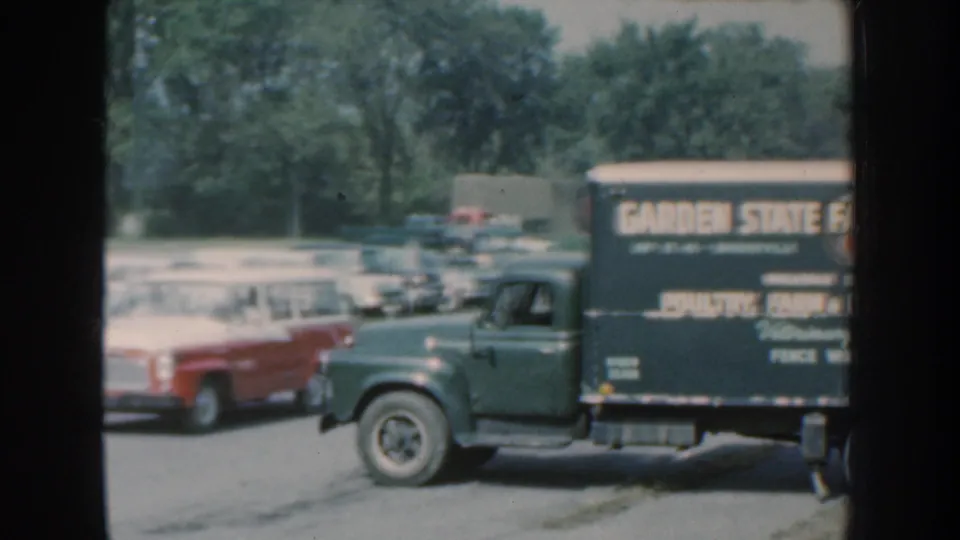 1959 Nebraska roadside frame showing a green commercial truck with lettering beside a red car and rows of parked vehicles.