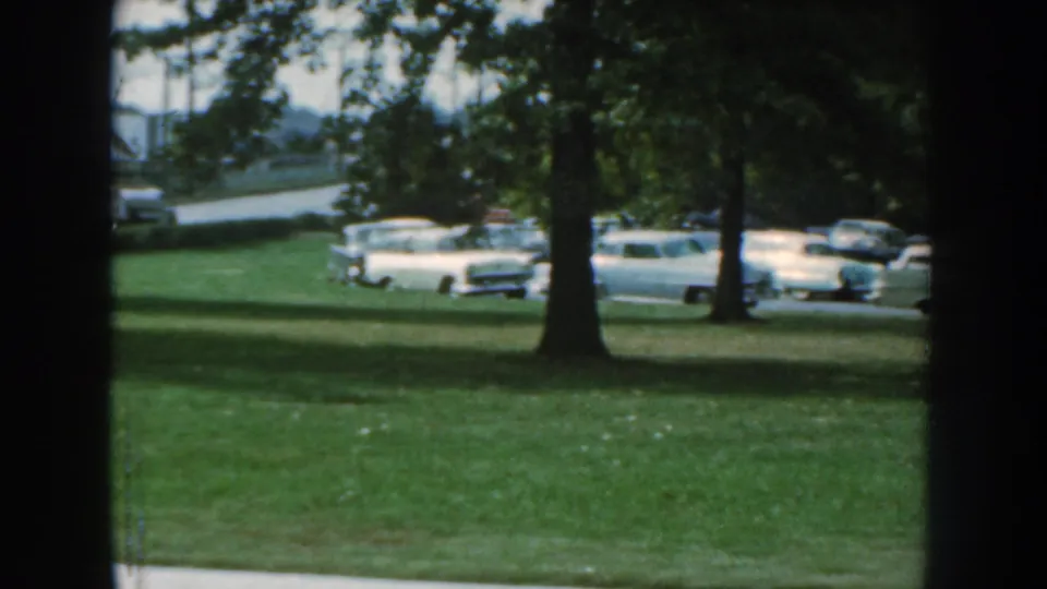 1959 Nebraska 8mm frame showing trees, grass, and parked mid-century automobiles near the exposition grounds.