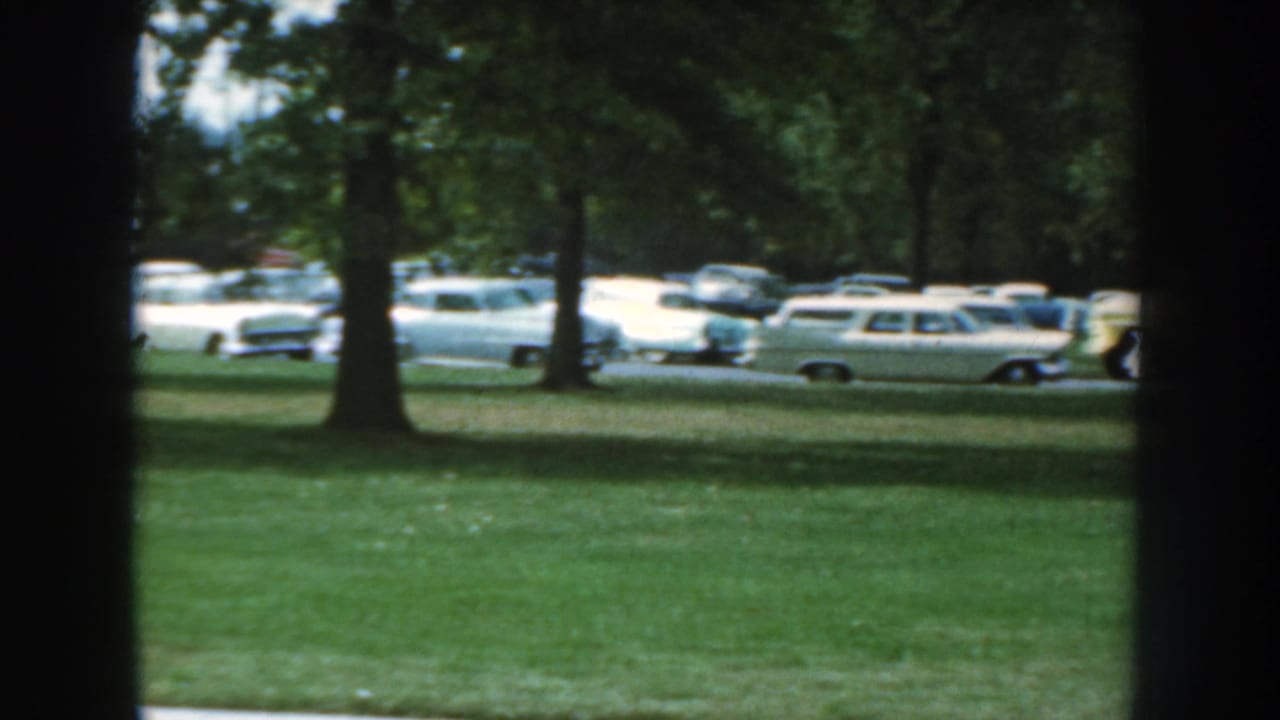1959 Nebraska home-movie view of parked cars and station wagons along a tree-lined street, framed by a dark interior edge.