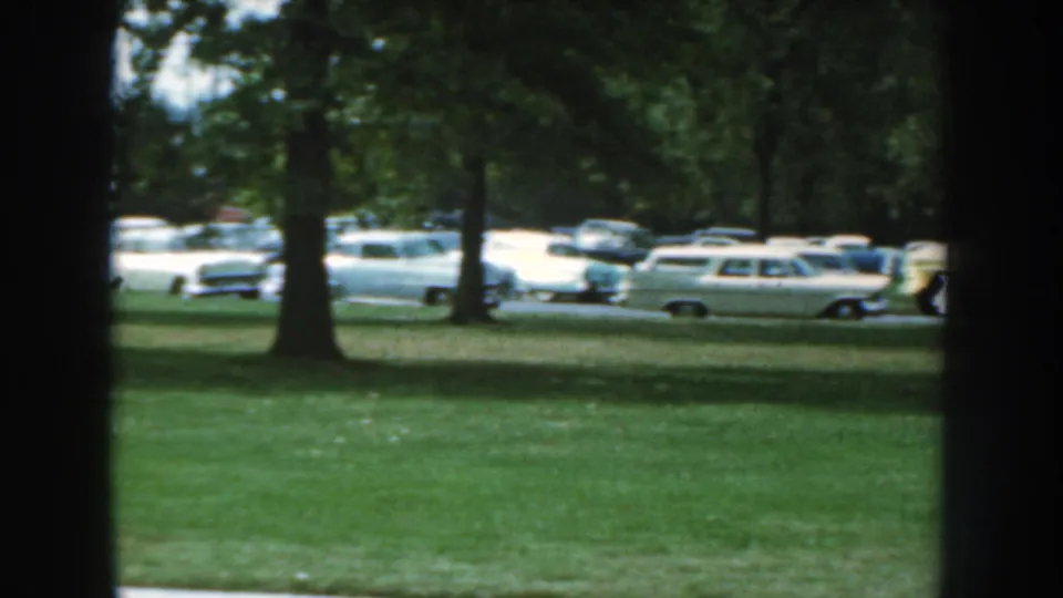1959 Nebraska home-movie view of parked cars and station wagons along a tree-lined street, framed by a dark interior edge.